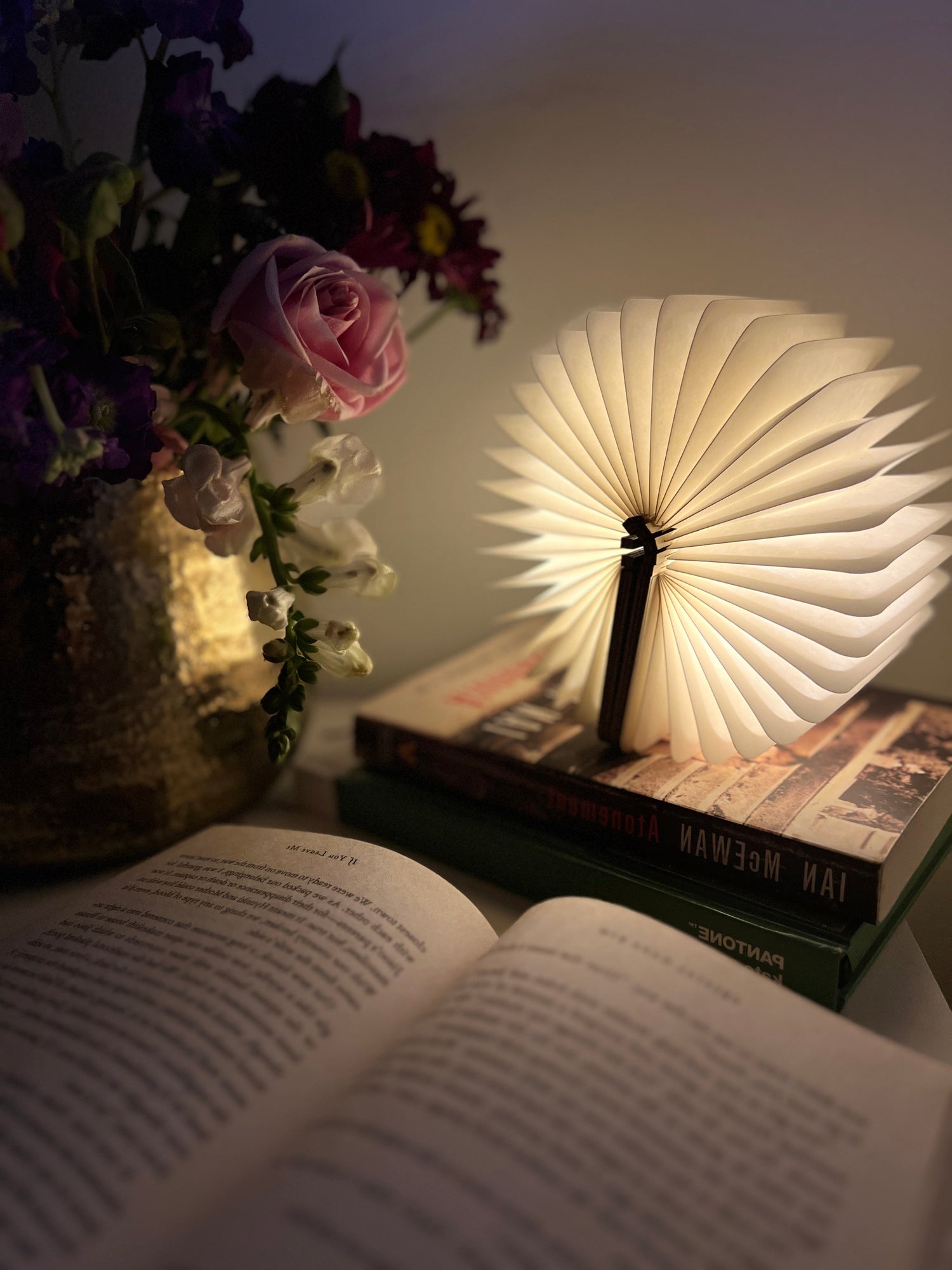A glowing book shaped lamp with fanned-out pages sits on top of two stacked books beside a golden vase of fresh flowers. An open book lies in the foreground, creating a warm and cozy reading nook ambiance.